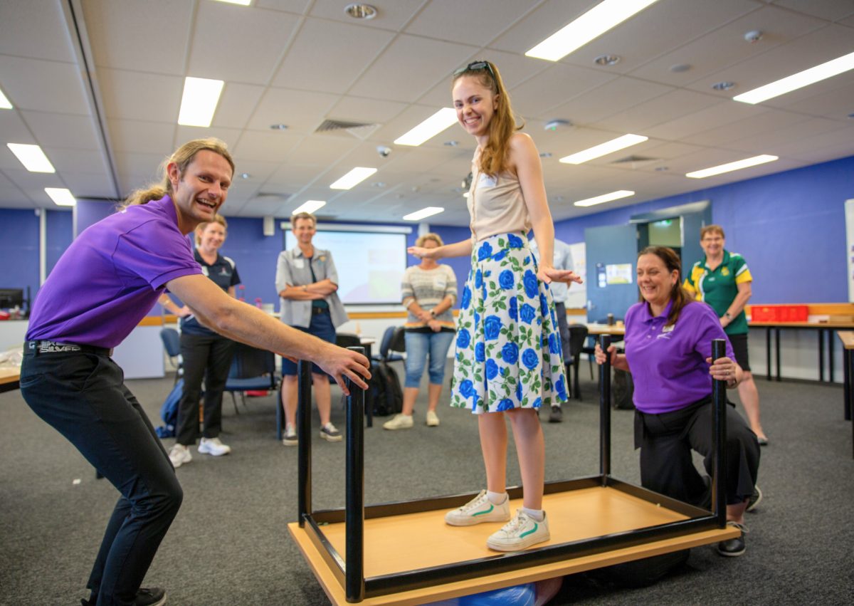 person balancing on an upside down table with two people helping