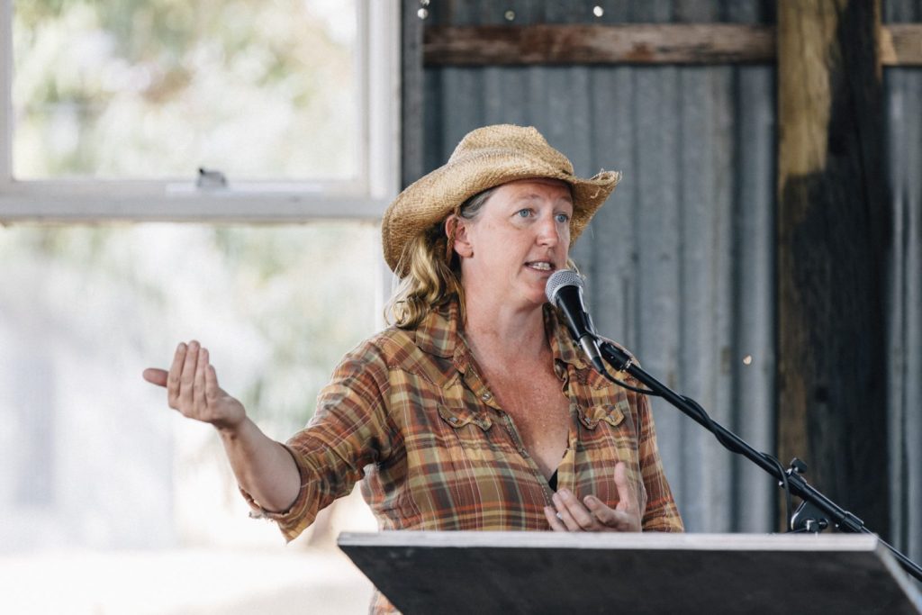 A woman in straw cowboy style hat and checked shirt speaks at a podium,