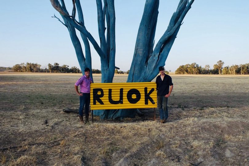 Two young men stand by a sign saying R U OK in front of a tree painted blue