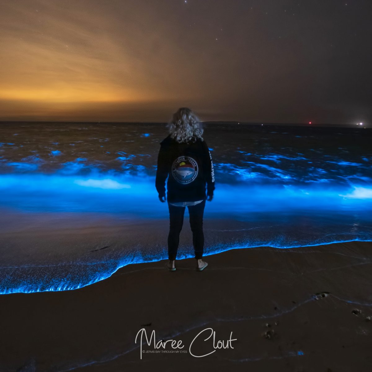 A woman with blonde curly hair is standing in front of the glowing waves at sunset. 