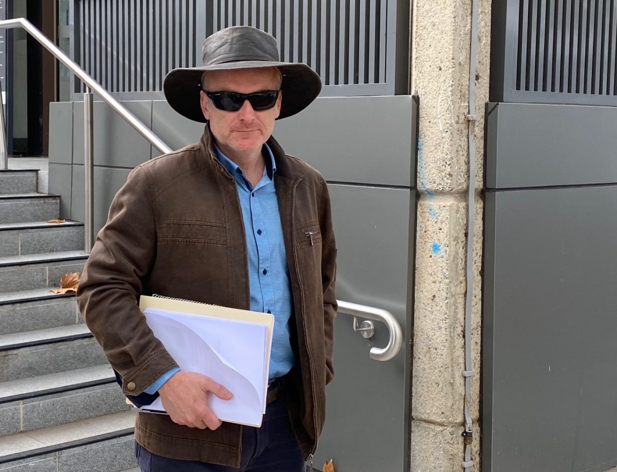 A man in sunglasses and wearing a hat, and holding documents, outside a building