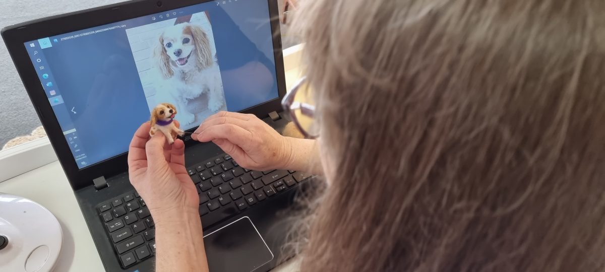 Woman looking at a pictyure of a dog on a laptop and crafting a miniature dog from felt