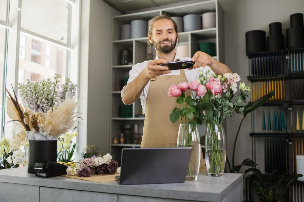 Small business owner in an apron takes photos of a vase of flowers for his social media pages