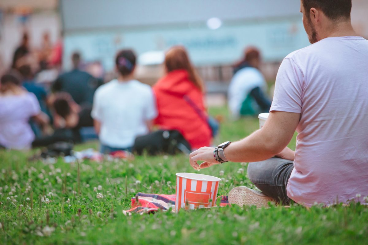 man sitting on the blanket eating snacks in front of big screen on open air cinema at public city park