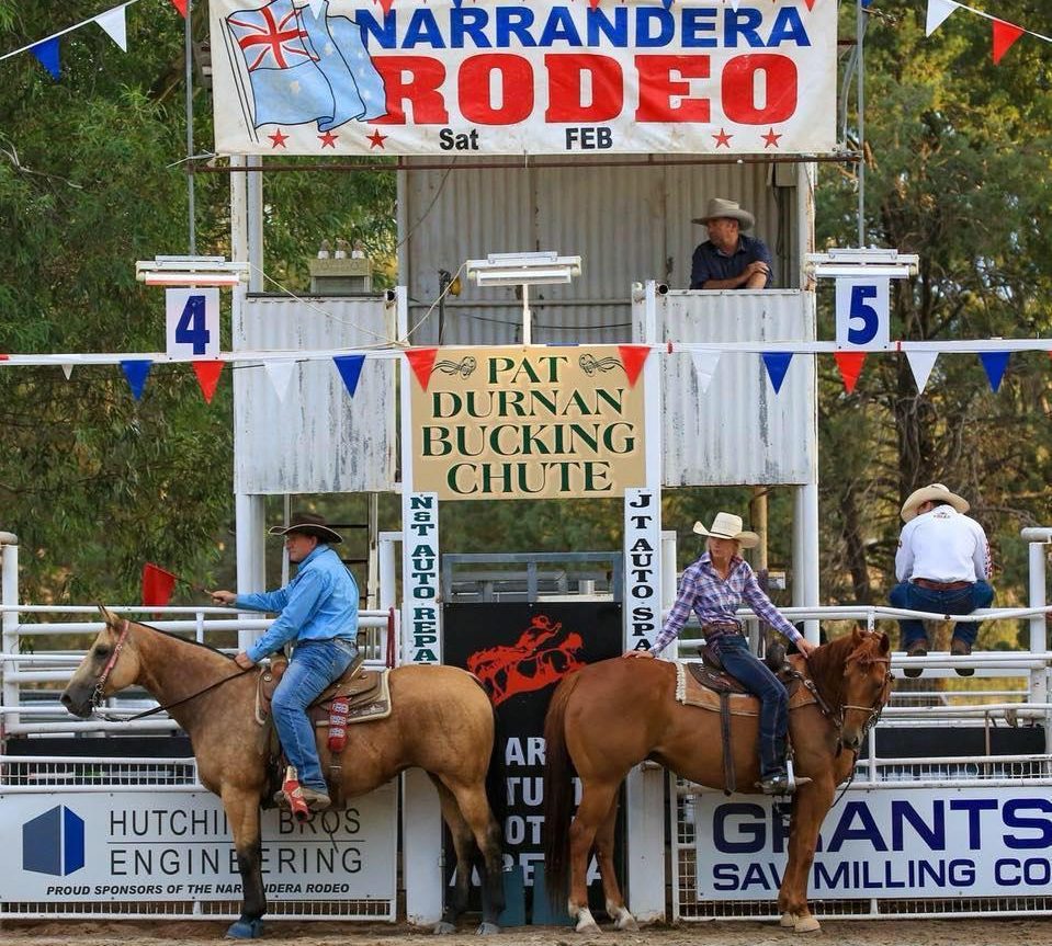 Two people on horses standing back to back under a sign that reads Narrandera Rodeo