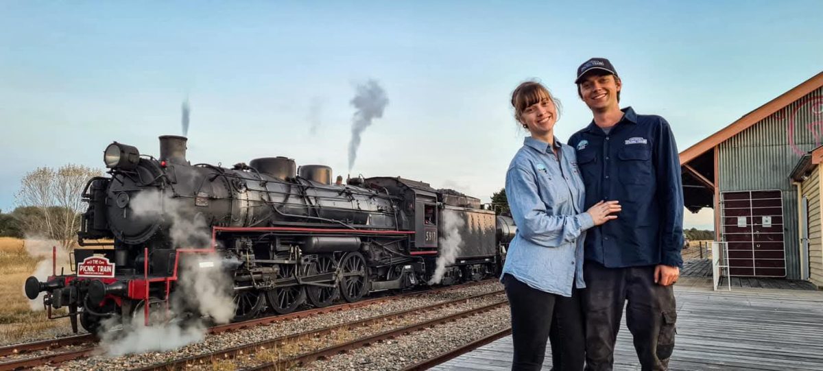 a woman and a man at a train station, with a steam train departing