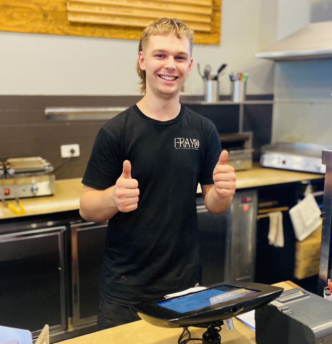 A young man standing in a cafe kitchen and giving a thumbs-up