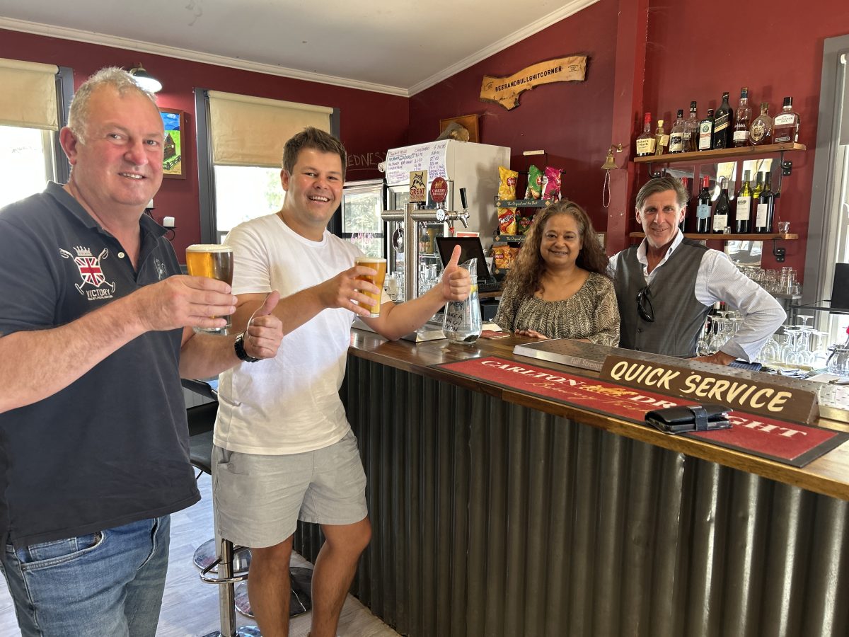 Two men drinking beer at a bar with owners behind the bar