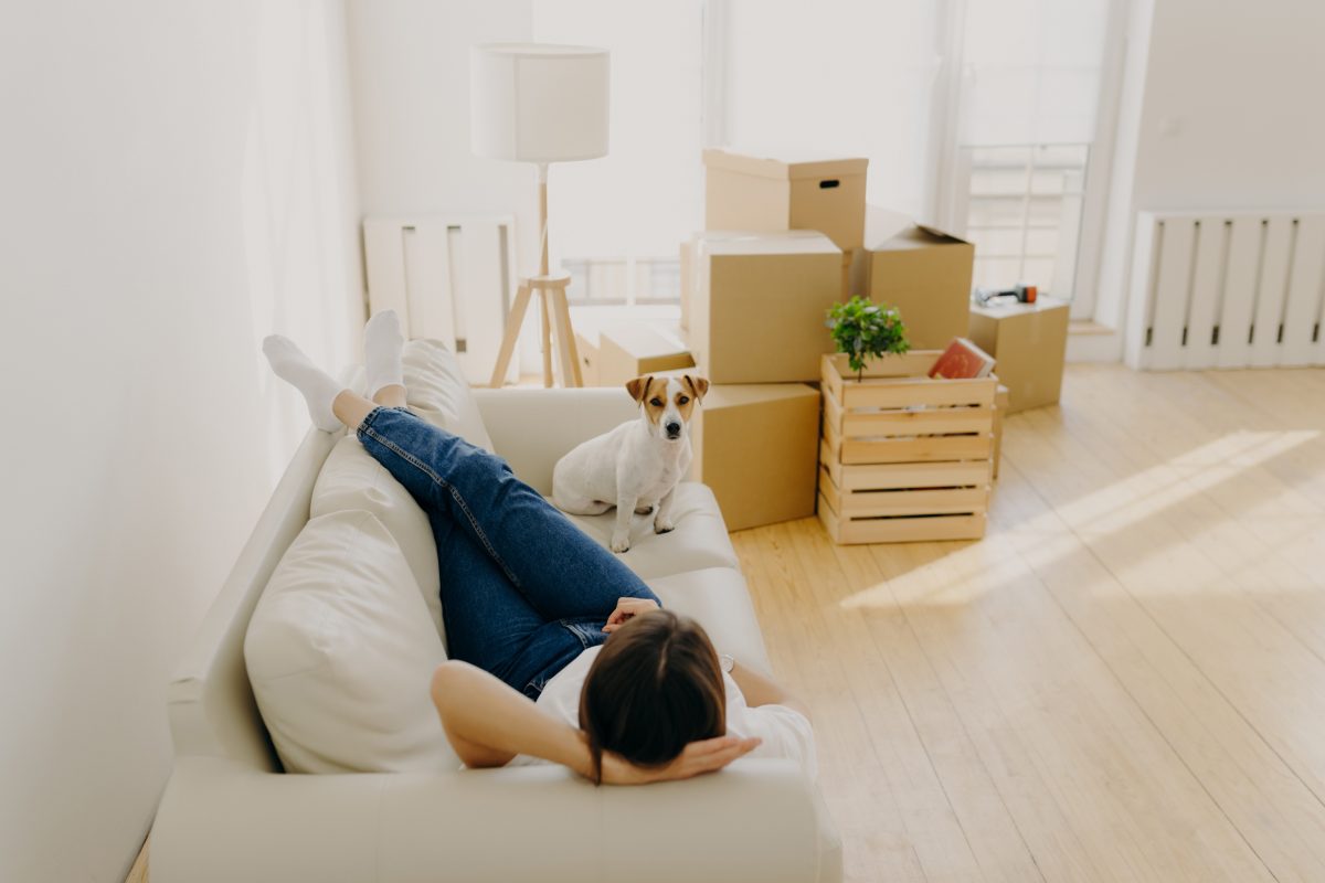Woman rests on sofa with pet dog, surrounded in boxes