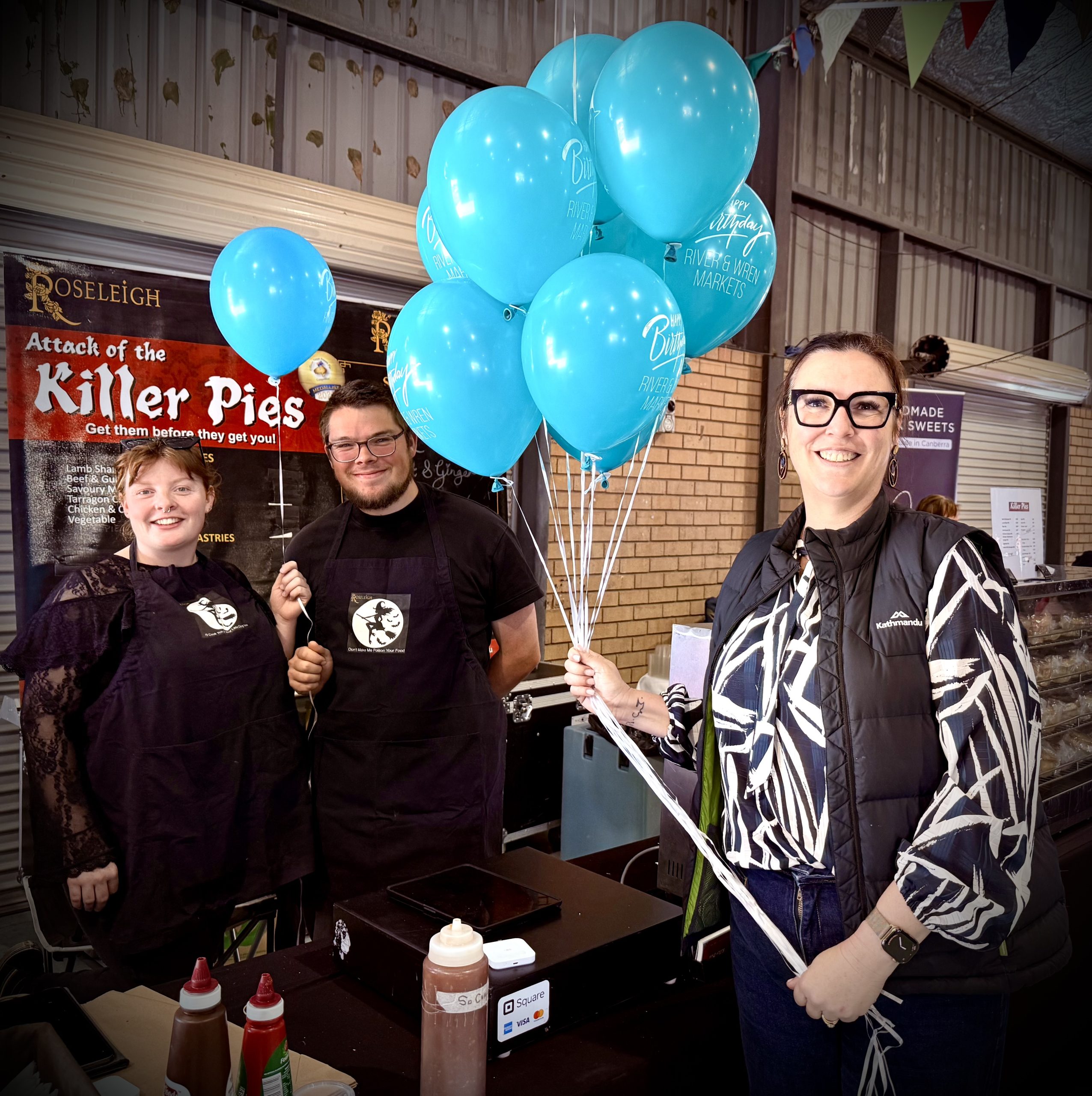three people with balloons at a market pie stall