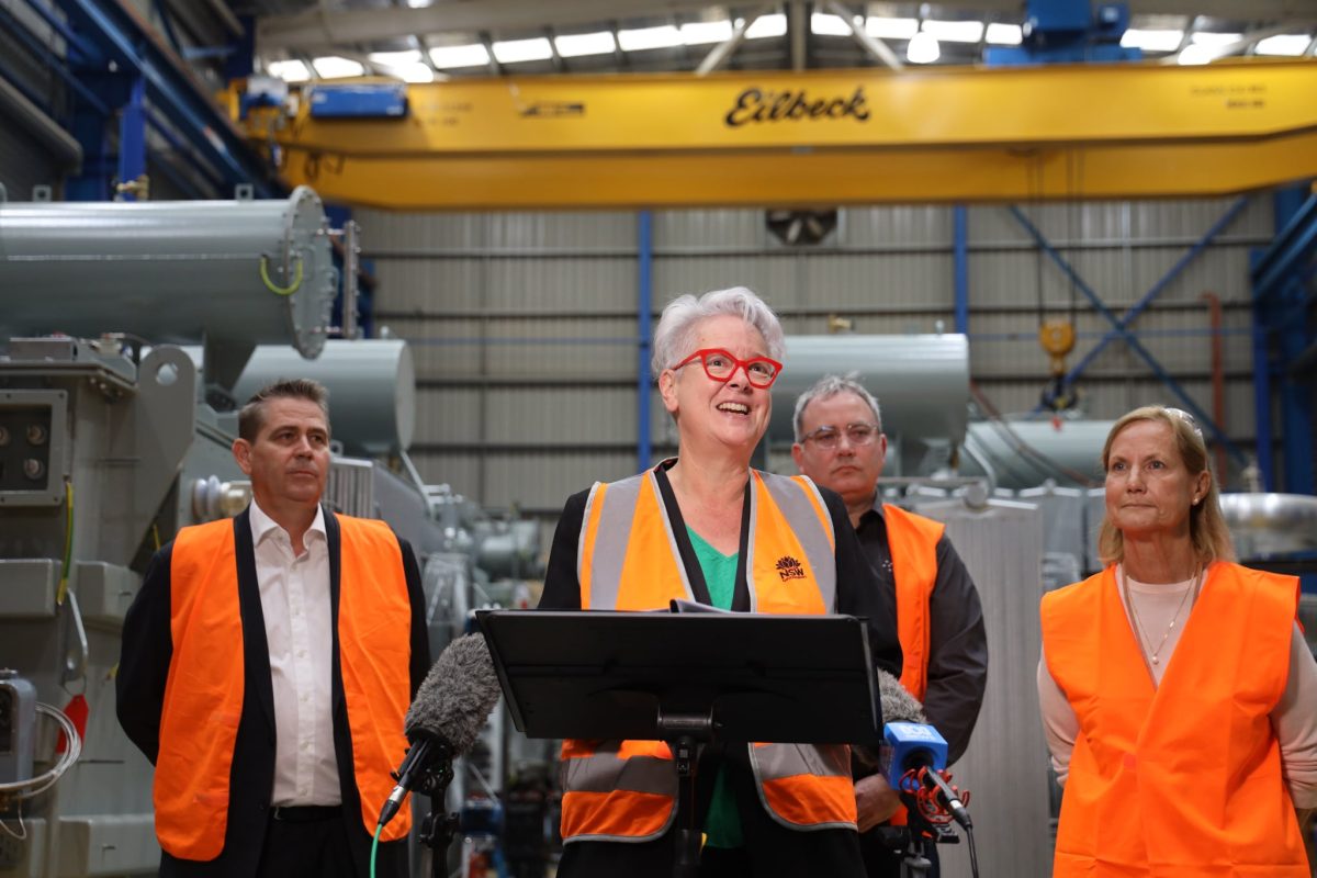 woman with red glasses and hi-vis vest and three other people holding a press conference in an industrial shed